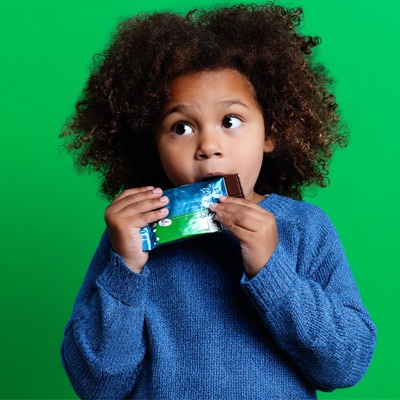 child-wearing-blue-sweater-eating-packaged-chocolate-bar-on-green-background